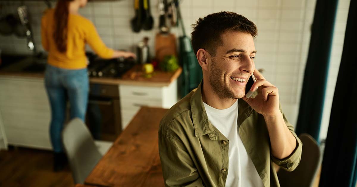 Man smiling while talking on phone with partner in background of kitchen.