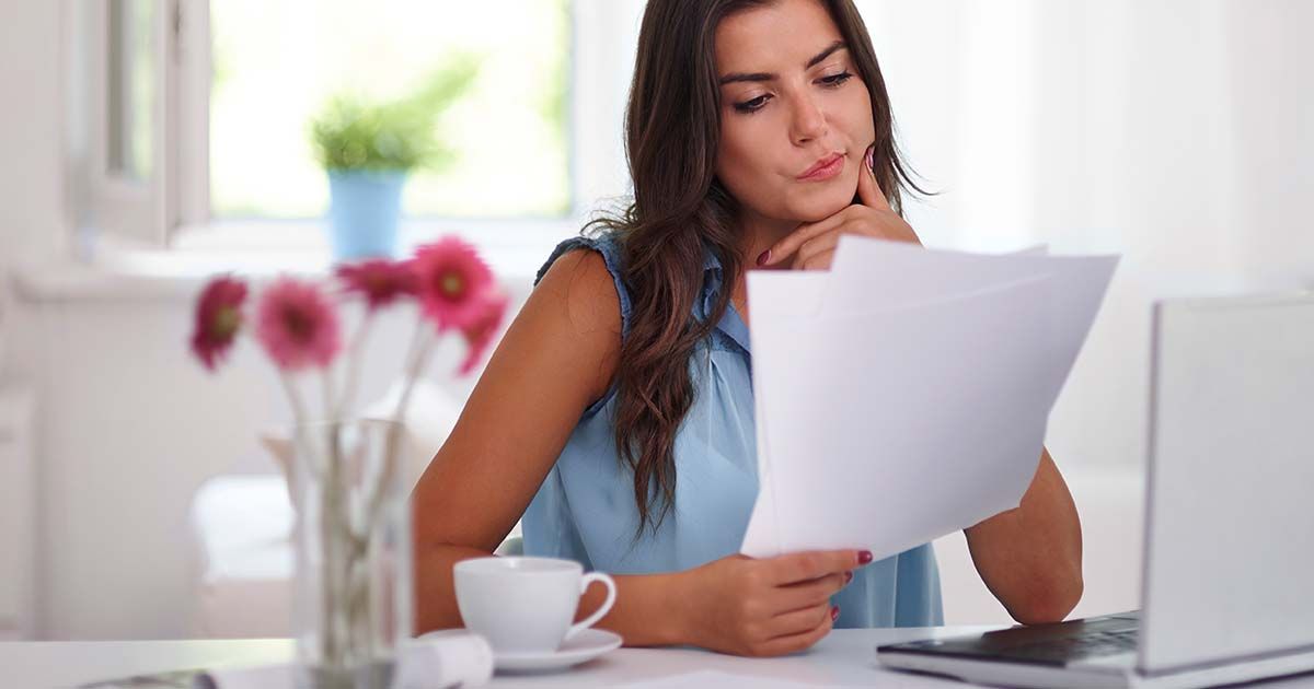 Woman looking at paperwork at dining table with laptop.