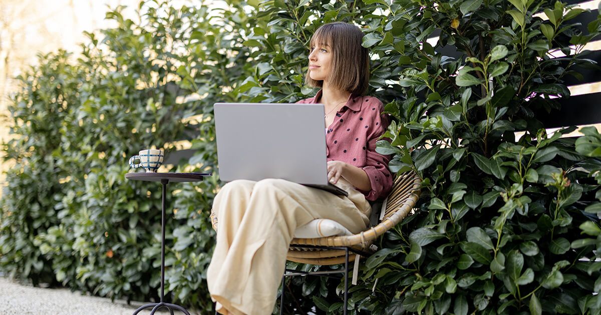 Woman sitting with laptop in backyard with bushed, privacy fence.